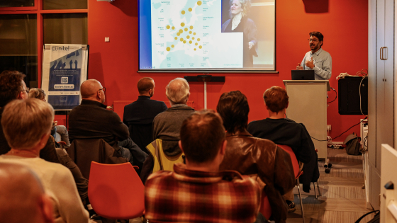 spreker in oranje zaal en aanwezigen die luisteren