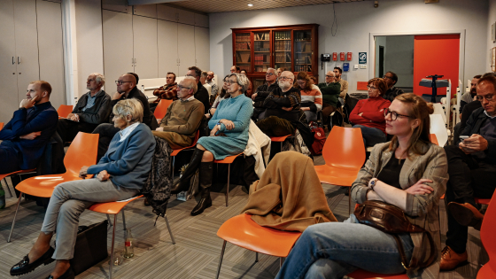 oranje zaal met aanwezigen die luisteren naar een lezing
