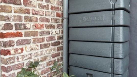 Grote donkergrijze regenton tegen de gevel. Ernaast staat een groene gieter en een bloempot met een plant