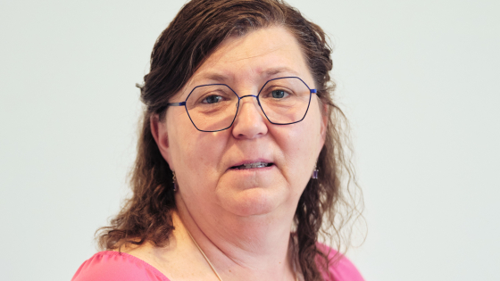 Dame met bruin haar en een bril. Ze draagt een roos t-shirt en een lange ketting