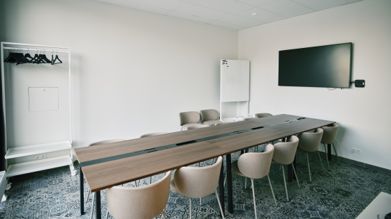 Ruime lichtrijke vergaderzaal. Er staat een lange bruine tafel met twaalf stoelen. Er hangt een televisiescherm aan de muur