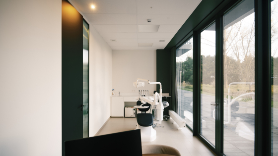Mooi modern tandartskabinet. Op de voorgrond een bureau met computerscherm, op de achtergrond de tandartsstoel. Het kabinet is lichtrijk dankzij de grote ramen.