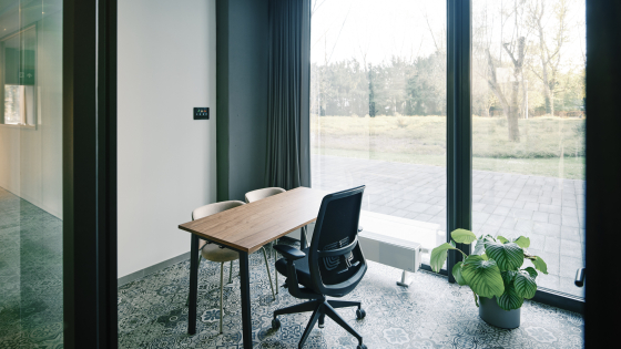 Kleine tafel met een bureaustoel. De ruimte heeft grote ramen, er staat een plant bij het raam