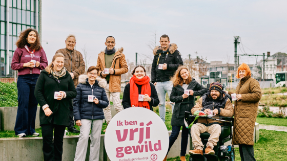 Groep van tien vrijwilligers poseert buiten. In het midden staat een rond logo met de woorden 'ik ben vrjij gewild. En gie?'