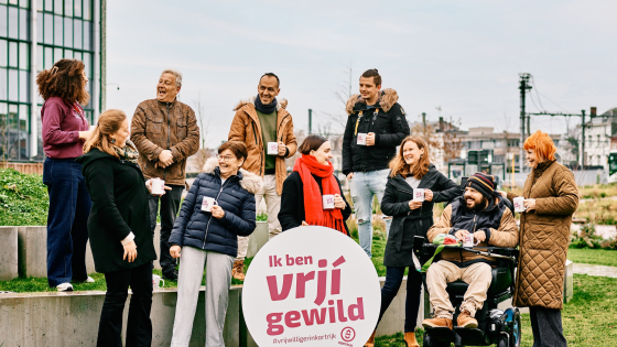 Groep van tien vrijwilligers poseert buiten. In het midden staat een rond logo met de woorden 'ik ben vrjij gewild. En gie?'
