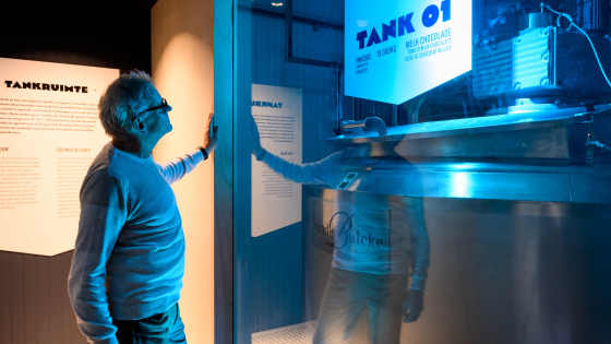 Een man bekijkt een grote glazen tank in een tentoonstellingsruimte. Op de tank staat de tekst "TANK 01" in blauwe letters, samen met technische informatie. De ruimte heeft een koel, blauw verlichtingsschema en aan de linkerkant is een informatief paneel zichtbaar met uitleg over tankprocessen.