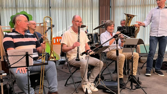 Groepsfoto. De leden van de band zitten op stoelen en hebben hun instrumenten vast: een schuiftrompet, trombone, fagot en gewone trompet