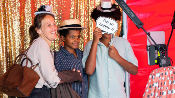 Een vrouw en twee jongens staan in een photobooth. Ze dragen leuke hoedjes en andere attributen voor op de foto
