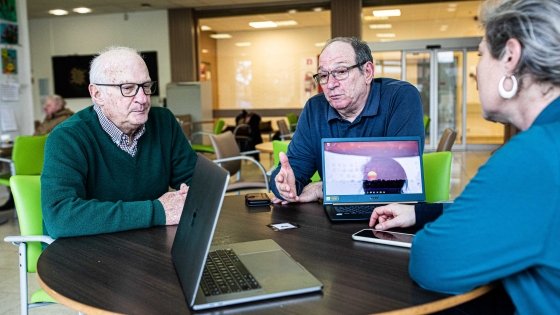 drie mensen zitten te praten aan een tafel, er staat twee computers op de tafel