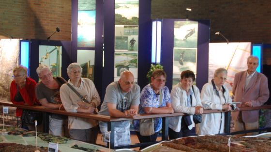 Een tiental leden van de Okra zijn in een museum, ze kijken naar een schaalmodel van een stad