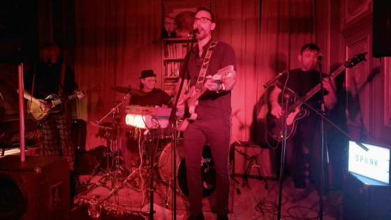 De leadzanger/gitarist staat op het podium met 2 bassisten en een drummer. Het podium baadt in rood licht