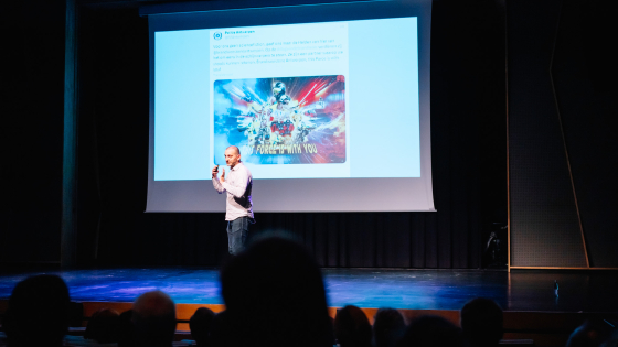 spreker digitaal jaaroverzicht 2023 op het podium van concertstudio Track Track met projectie op het scherm van het digitale jaaroverzicht 2023