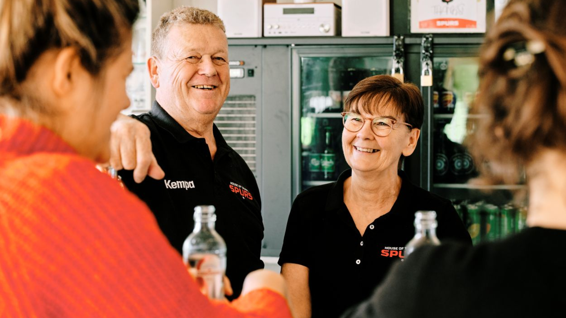 Carine en Lieven staan achter de toog. Ze dragen beiden een zwarte polo. Ze lachen vrolijk