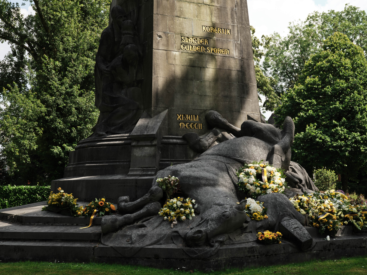 Foto van de sokkel van het Groeningemonument, close-up op het gevallen paard, omringd door bloemenkransen.