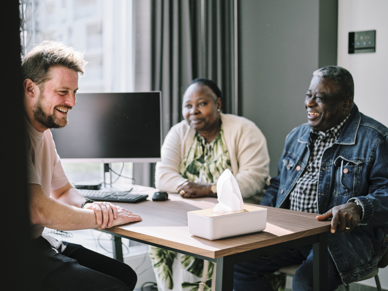 3 mensen zitten aan tafel en voeren een gesprek. Op de tafel staat een computerscherm en een doos zakdoeken
