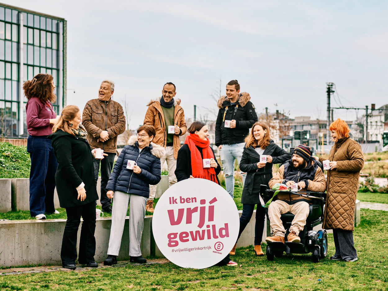 Groep van tien vrijwilligers poseert buiten. In het midden staat een rond logo met de woorden 'ik ben vrjij gewild. En gie?'