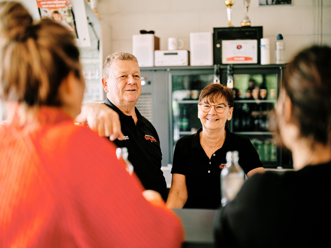 Carine en Lieven staan achter de bar, ze praten met twee vrouwen