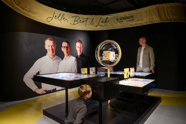 Een tentoonstellingsruimte met een groot portret van drie mannen naast een ronde vitrine met strips. Een jongen knielt op de grond en kijkt naar een van de uitgestalde items, terwijl een man in een beige jasje naast de vitrine staat. Bovenaan hangt een gele banner met de namen "Jeff, Bert & Luc".