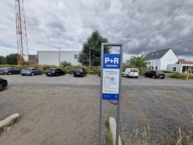 foto van een parking waarop een vijftal auto's geparkeerd staat. Op de voorgrond staat een infobord over de parking en de gratis bus.