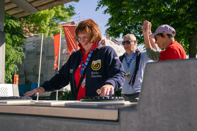 Een vrouw staat aan een dj-tafel. Achter haar staan 2 mannen te dansen. Ze staan in de buitenlucht