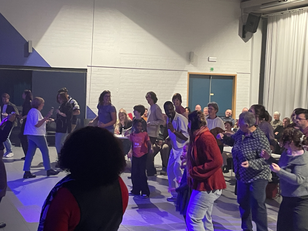 Een groep mensen staat in een cirkel te dansen. Een zwarte man in witte kleren zingt in de micro.  Op de achtergrond zitten nog wat mensen aan tafels, zij durven wellicht niet dansen