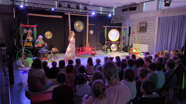 Een klas jonge kinderen kijkt naar een voorstelling. Op sc&egrave;ne zingt een jongedame in een witte jurk met rode bloemen. Achter haar speelt een muzikant op de trekzak. Op de sc&egrave;ne staat nog een klein rood brugje, een grote metalen gong en wat planten en kommen, allemaal in Oosterse sferen.