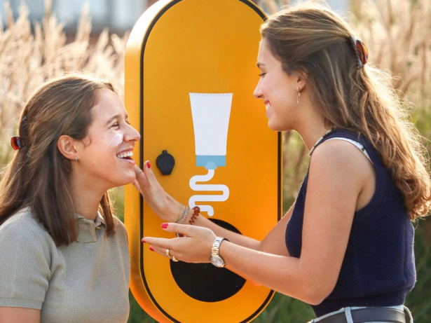 Twee jonge vrouwen bij een SMOT-pot. De ene vrouw smeert het gezicht van de andere in met zonnecr&egrave;me.