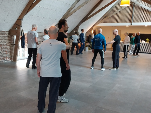 Verschillende duo's staan in een zaal. Ze hebben sportieve kleren aan voor deze activiteit